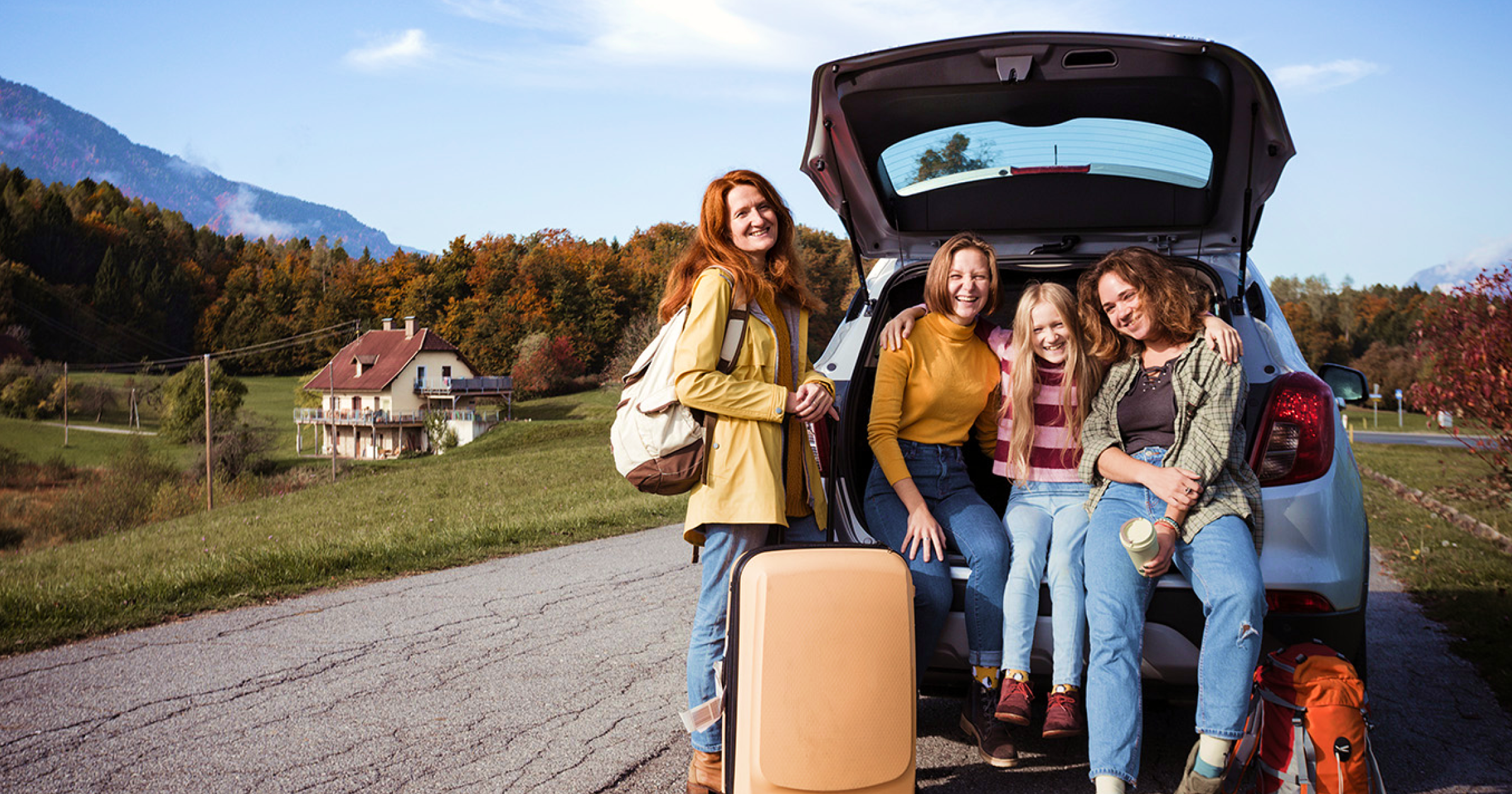 A happy group of girls, standing by their car with luggage, enjoys a scenic fall destination, surrounded by colorful autumn trees and mountains.