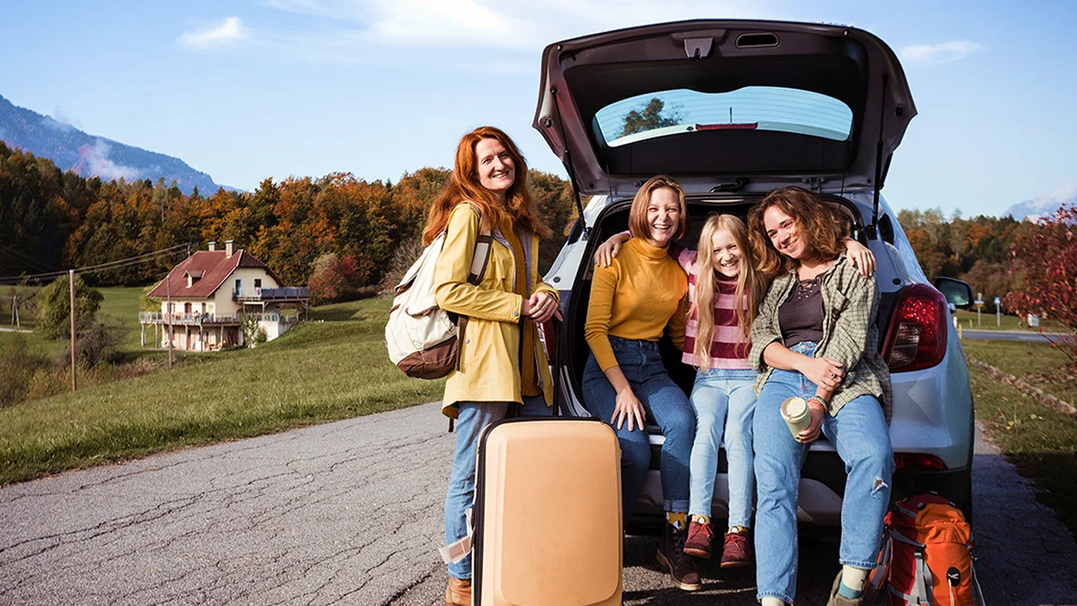 A happy group of girls, standing by their car with luggage, enjoys a scenic fall destination, surrounded by colorful autumn trees and mountains.